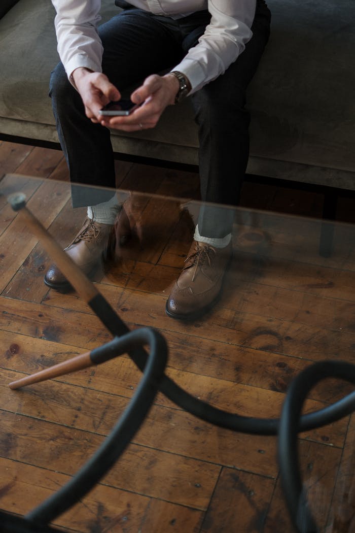 Man sitting on a sofa using a smartphone, seen through glass table indoors.